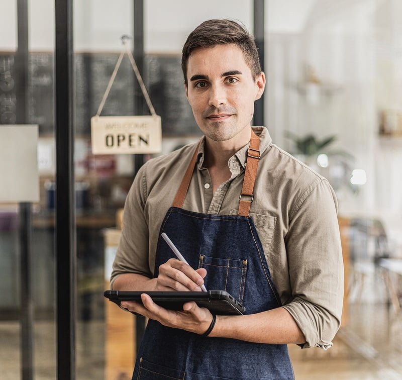 A handsome man is in a cafe, he's standing holding a mockup tablet for a welcome message or a menu, he's a cafe worker wearing an apron and serving customers. Cafe service concept.