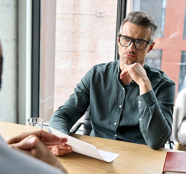 Serious focused mature businessman employer hr or client holding cv or contract listening manager, applicant during job interview, negotiations office meeting thinking or making business decision.